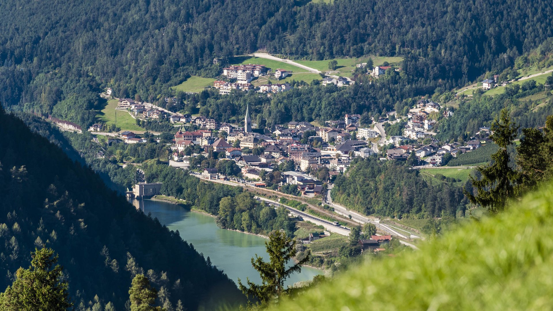 Mühlbach im Pustertal Das Bild zeigt eine kleine Stadt, die sich in einem grünen Tal erstreckt, umgeben von dicht bewaldeten Hügeln. In der Mitte der Stadt ragt ein Kirchturm heraus, und im Vordergrund ist ein See zu sehen, an dessen Ufer sich ein Gebäude befindet.