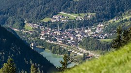 Mühlbach im Pustertal Das Bild zeigt eine kleine Stadt, die sich in einem grünen Tal erstreckt, umgeben von dicht bewaldeten Hügeln. In der Mitte der Stadt ragt ein Kirchturm heraus, und im Vordergrund ist ein See zu sehen, an dessen Ufer sich ein Gebäude befindet.