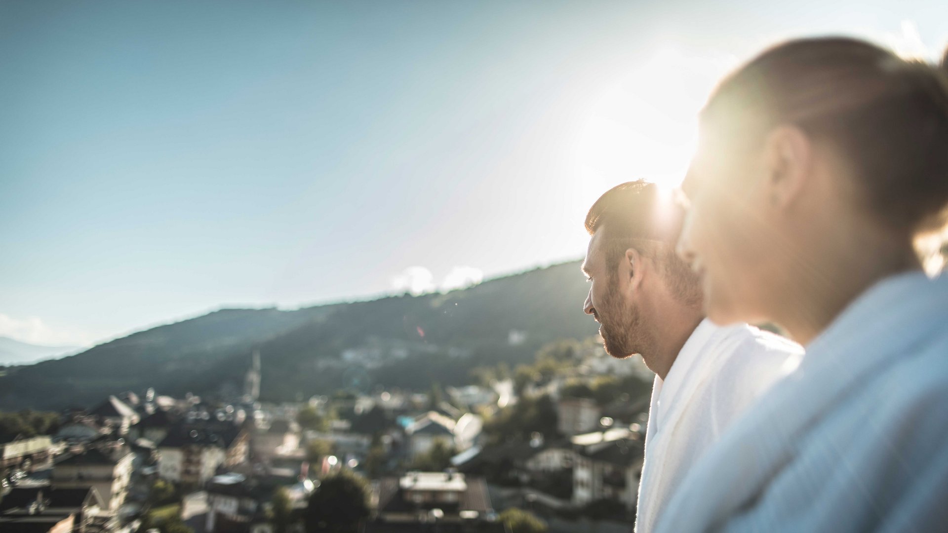 Mühlbach im Pustertal Das Bild zeigt zwei Personen, die von hinten in die Ferne schauen, während die Sonne über einem Berg im Hintergrund scheint. Die Personen tragen weiße Bademäntel, und die Szenerie ist leicht unscharf, wodurch ein weiches, verträumtes Ambiente entsteht.