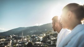 Mühlbach im Pustertal Das Bild zeigt zwei Personen, die von hinten in die Ferne schauen, während die Sonne über einem Berg im Hintergrund scheint. Die Personen tragen weiße Bademäntel, und die Szenerie ist leicht unscharf, wodurch ein weiches, verträumtes Ambiente entsteht.