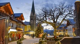 Advent in Terenten The picture shows a festively decorated square with a church in the background. A large tree wrapped in lights and a decorated Christmas tree stand amid houses, which are also adorned with string lights, while snow covers the ground.