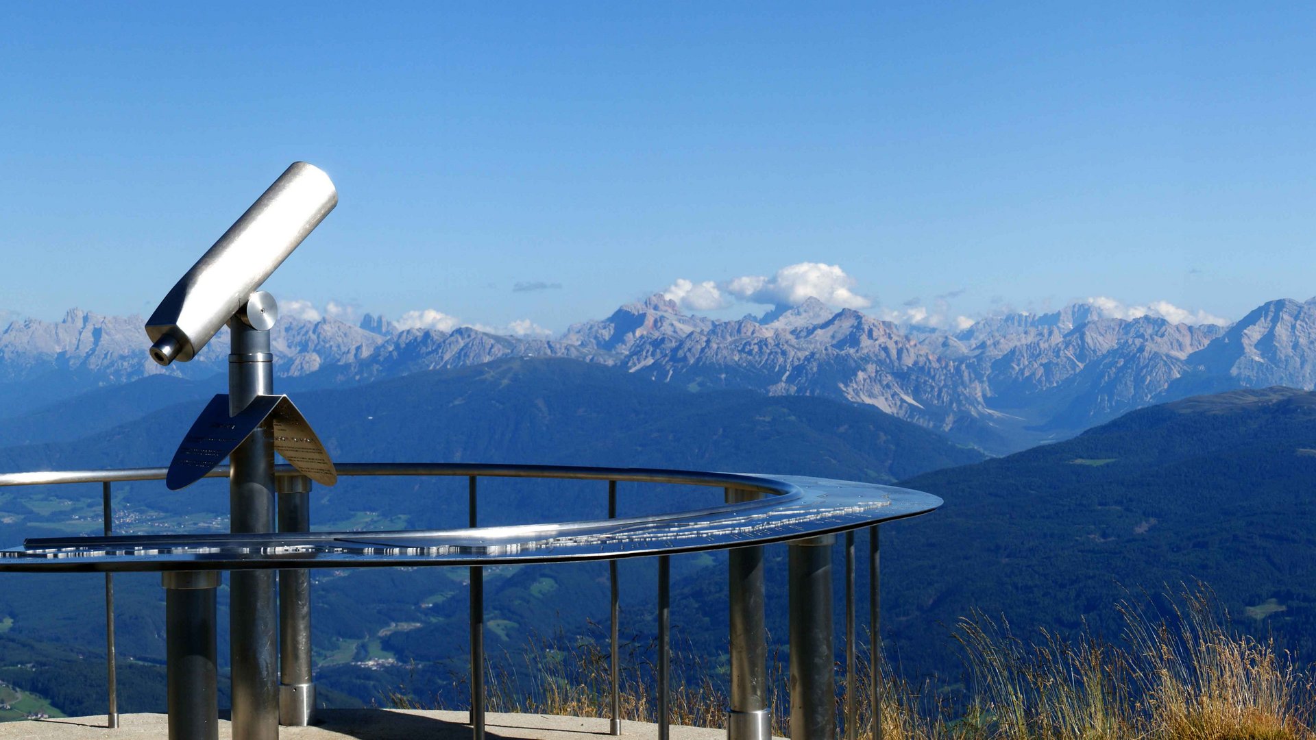 The Gitschberg panoramic platform in Meransen, Italy The picture shows a panoramic viewing platform on a mountain peak, equipped with a permanently installed telescope. In the background, a wide valley stretches out, surrounded by numerous mountain ranges and peaks, under a clear, blue sky.