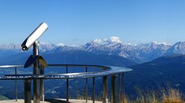 The Gitschberg panoramic platform in Meransen, Italy The picture shows a panoramic viewing platform on a mountain peak, equipped with a permanently installed telescope. In the background, a wide valley stretches out, surrounded by numerous mountain ranges and peaks, under a clear, blue sky.