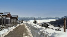 The mountain village of Meransen The picture shows a narrow asphalt road running through a snowy landscape. On the left side of the road, there are wooden houses and a fence, while in the background a dense layer of clouds covers the valleys and tall snow-covered mountains can be seen under a clear blue sky.