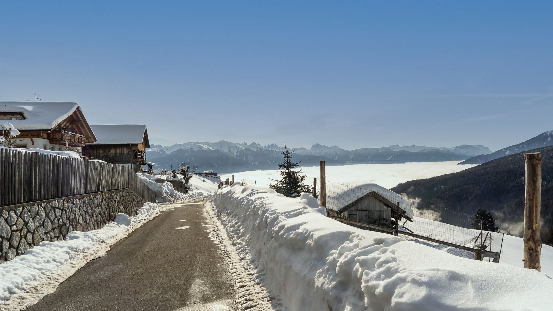 The mountain village of Meransen The picture shows a narrow asphalt road running through a snowy landscape. On the left side of the road, there are wooden houses and a fence, while in the background a dense layer of clouds covers the valleys and tall snow-covered mountains can be seen under a clear blue sky.