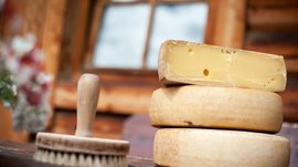 Fane Alm – South Tyrol’s most idyllic Alpine village The picture shows several cheese wheels, one of which is cut, allowing to see the yellow interior with some holes. Next to it lies a round brush on a wooden table, and in the background a window is visible out of focus.