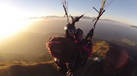 Paragliding v Jižním Tyrolsku Obrázek ukazuje dvě osoby, které během paraglidingu plují vzduchem, zatímco slunce je nízko na obloze a zalévá krajinu zlatavým světlem. V pozadí jsou vidět hory a rozsáhlá rovina, která se ztrácí v jemné mlze.