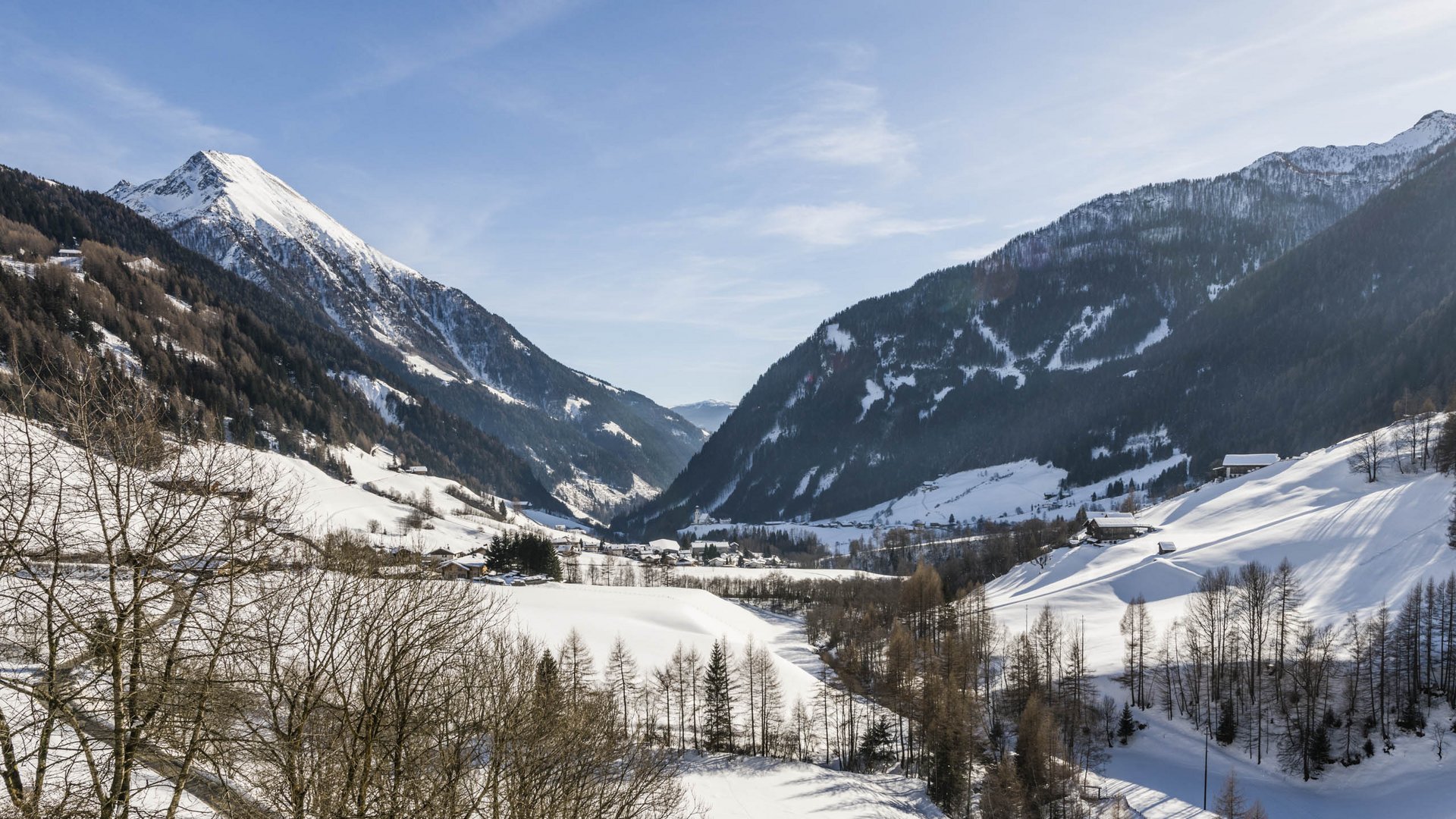 Pfunders: urig und idyllisch Das Bild zeigt eine winterliche Berglandschaft mit schneebedeckten Hängen und einem Tal in der Mitte. Die Sonne scheint, der Himmel ist klar und blau, und im Vordergrund sind kahle Bäume zu sehen. Die Berge im Hintergrund sind ebenfalls mit Schnee bedeckt, was eine friedliche und ruhige Atmosphäre vermittelt.