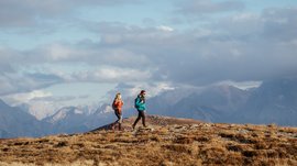 De mooiste wandelroutes in de Dolomieten Het beeld toont twee wandelaars, een man en een vrouw, die in warme kleding en met rugzakken over een met gras begroeid berglandschap lopen. Op de achtergrond zijn hoge, deels met wolken bedekte bergen te zien.