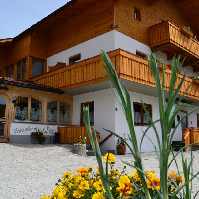 Selský šenk Heisla Dvoupodlažní budova s bílou fasádou a dřevěnými balkony, před kterou kvetou barevné květiny. Vchod je zdoben dekorativními okny a cedulí s nápisem "Häuslerhof".