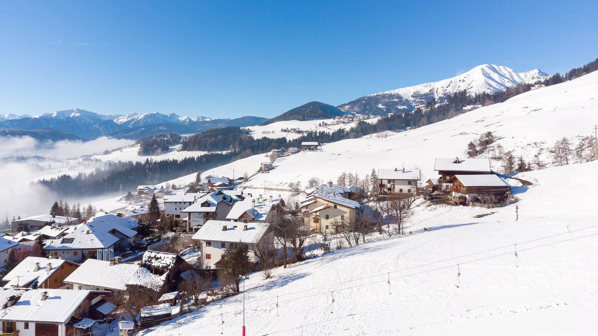 Skivakantie met kinderen in Zuid-Tirol/Terenten De afbeelding toont een besneeuwd dorp in de bergen, met met sneeuw bedekte daken en heuvels. Op de achtergrond zijn beboste heuvels en majestueuze bergen te zien onder een heldere, blauwe lucht.