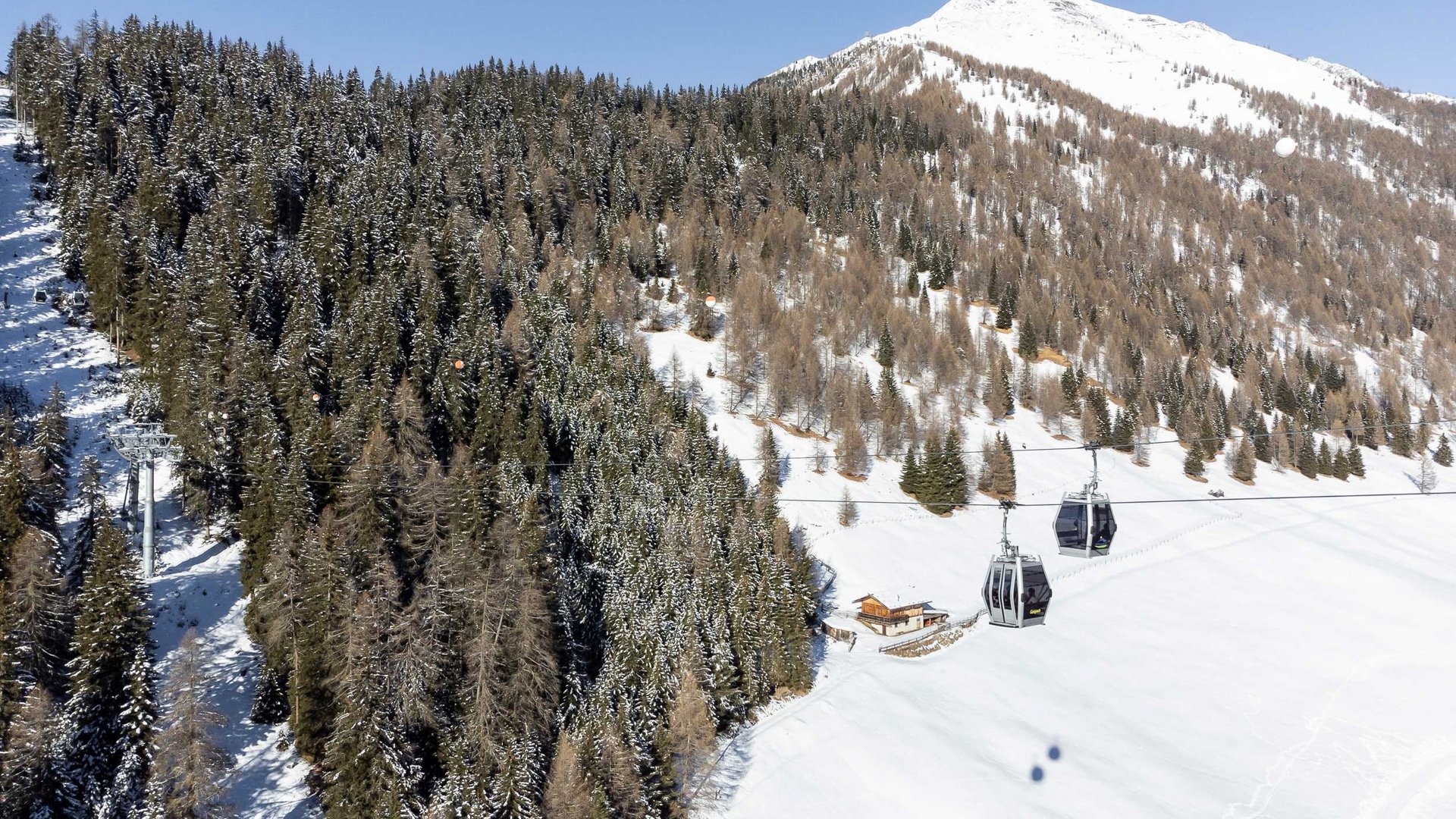 The Altfasstal valley, Meransen The picture shows a cable car with gondolas traveling over a snowy landscape. In the background, dense snow-covered forests and a high snow-covered mountain peak can be seen. At the foot of the slope, there is a small hut. The sky is clear and blue.