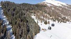 The Altfasstal valley, Meransen The picture shows a cable car with gondolas traveling over a snowy landscape. In the background, dense snow-covered forests and a high snow-covered mountain peak can be seen. At the foot of the slope, there is a small hut. The sky is clear and blue.