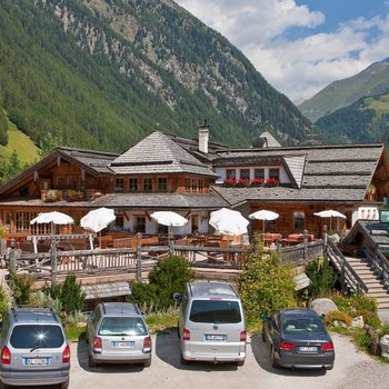 Südtiroler Almkost Das Bild zeigt ein großes, rustikales Berghotel mit Holz- und Steindach, umgeben von grünen Bergen und Wäldern. Vor dem Hotel befinden sich eine Terrasse mit weißen Sonnenschirmen und mehreren geparkten Autos, und ein Schild weist auf eine Pizzeria, ein Café und eine Skischule hin.