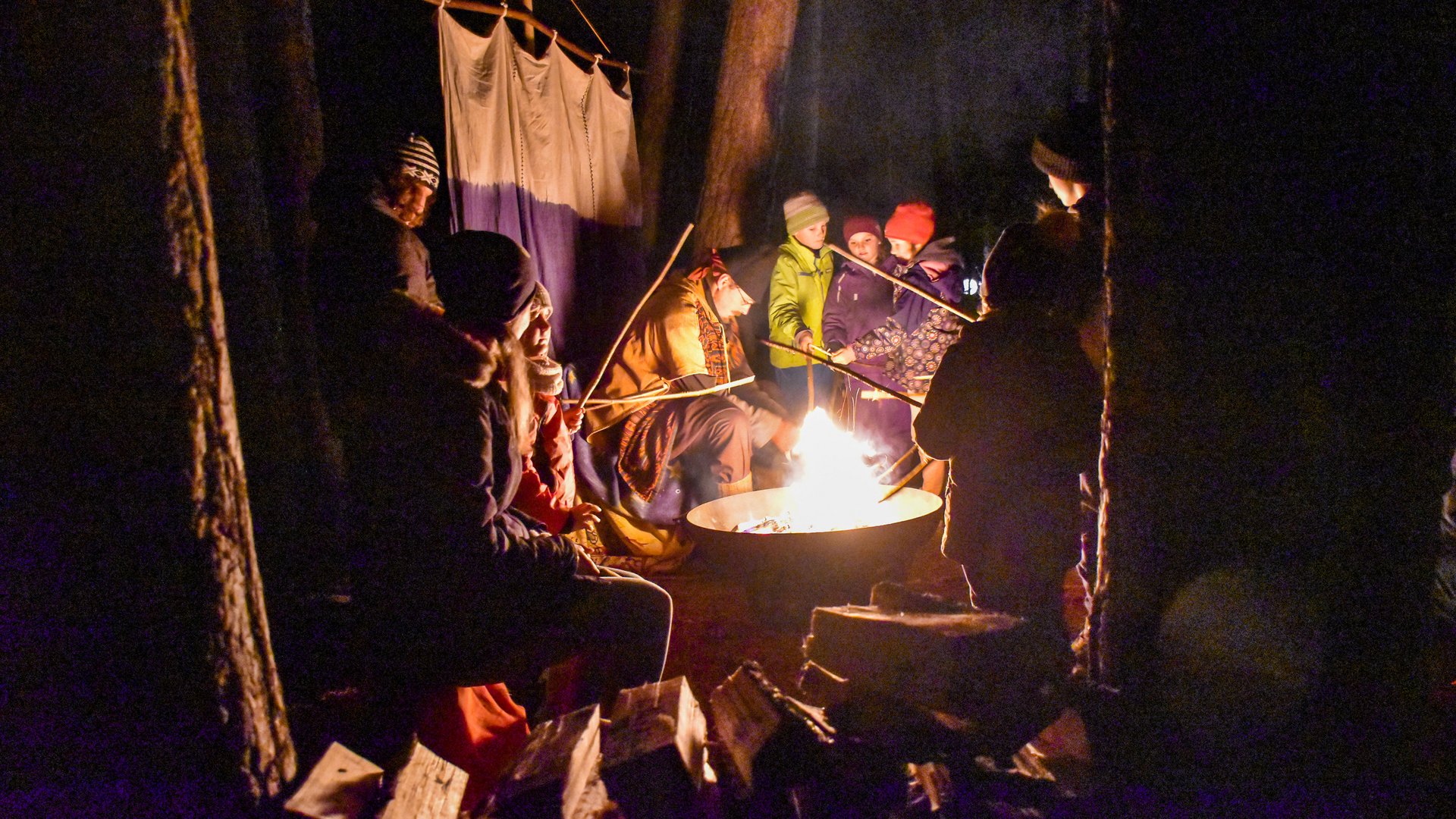 Advent in Terenten The picture shows a group of people gathered around a campfire. They are holding long sticks into the fire, presumably to roast something, and are dressed warmly, while the firelight illuminates their faces in the darkness.