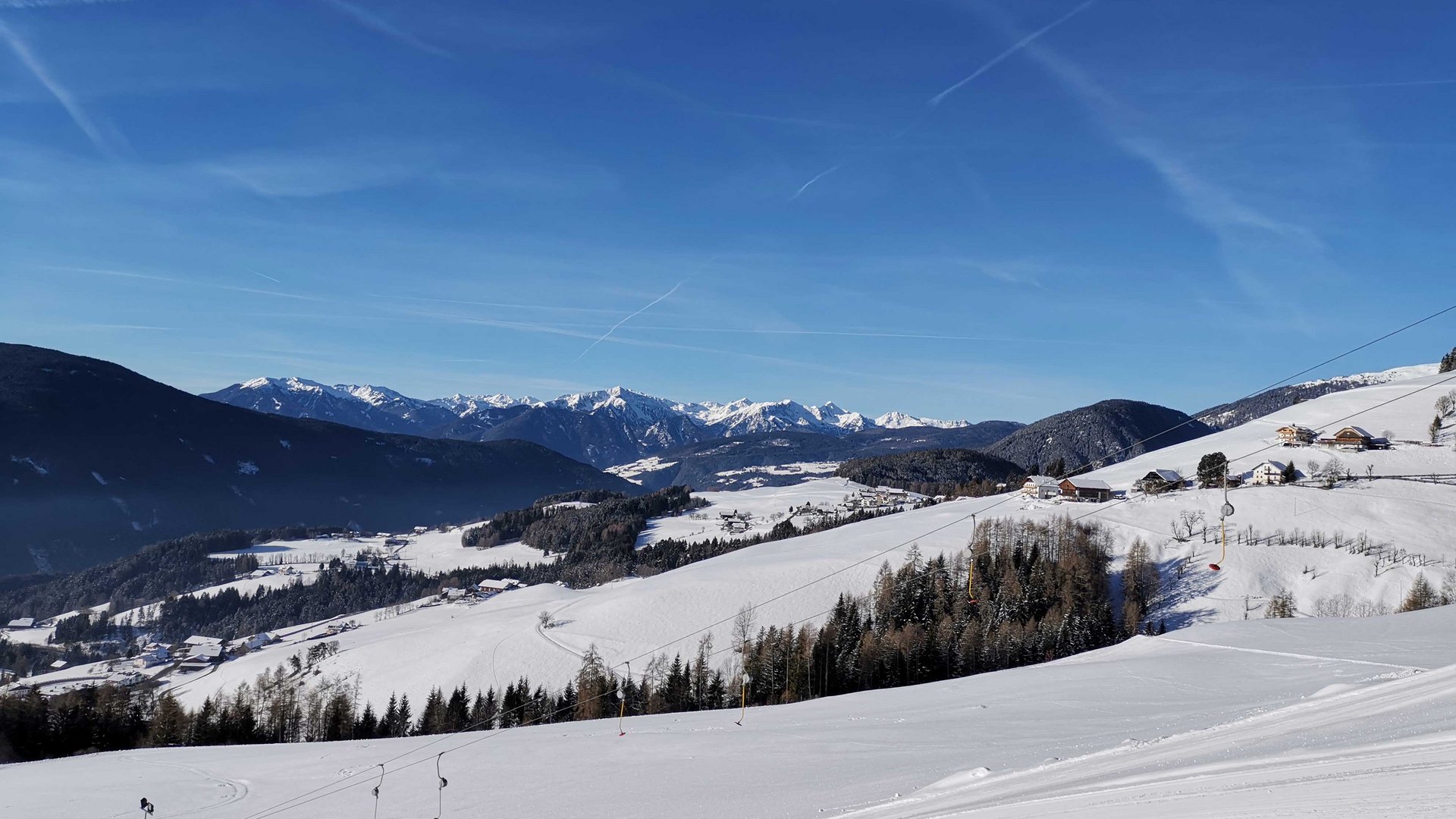 Skivakantie met kinderen in Zuid-Tirol/Terenten De afbeelding toont een met sneeuw bedekt berglandschap met een heldere blauwe lucht. Op de voorgrond zijn skiliften en hellingen te zien, terwijl zich op de achtergrond verschillende bergketens uitstrekken.
