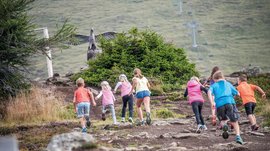 Sluneční park Gitschberg Obrázek ukazuje skupinu dětí, které stoupají po kamenité cestě. V pozadí je vidět velká socha orla s roztaženými křídly, skrytá za keřem.