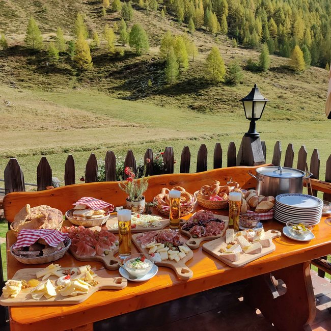 Pranter Stadelhütte Das Bild zeigt einen rustikalen Holztisch im Freien, der mit einer Vielzahl von Speisen gedeckt ist, darunter Käse, Brot, Aufschnitt und Brezeln. Im Hintergrund ist eine malerische Landschaft mit grünen Hügeln und vereinzelten Bäumen zu sehen.