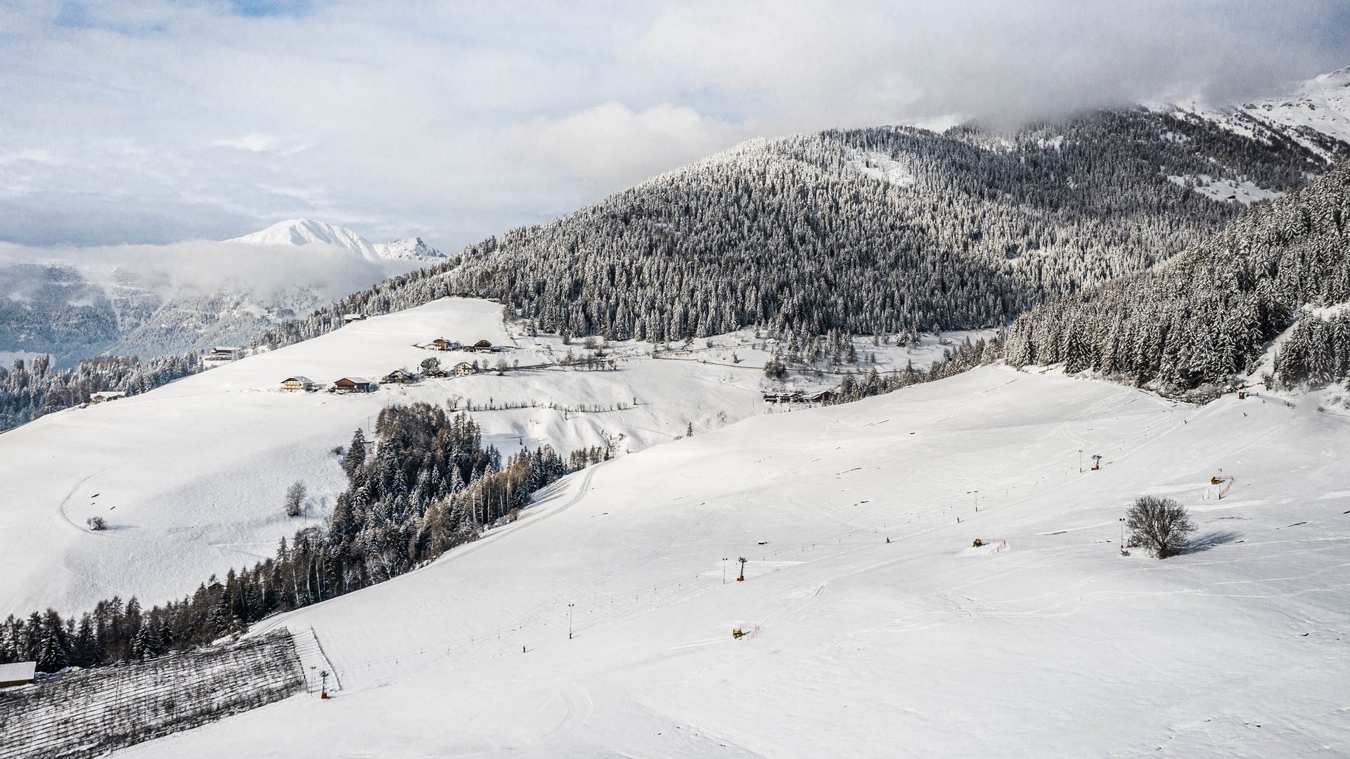 Skivakantie met kinderen in Zuid-Tirol/Terenten De afbeelding toont een uitgestrekt, met sneeuw bedekt landschap met zachte heuvels en een dicht bos op de achtergrond. Op de heuvels zijn verspreide huizen en skiliften te zien, terwijl de bergen op de achtergrond gedeeltelijk door wolken worden bedekt.
