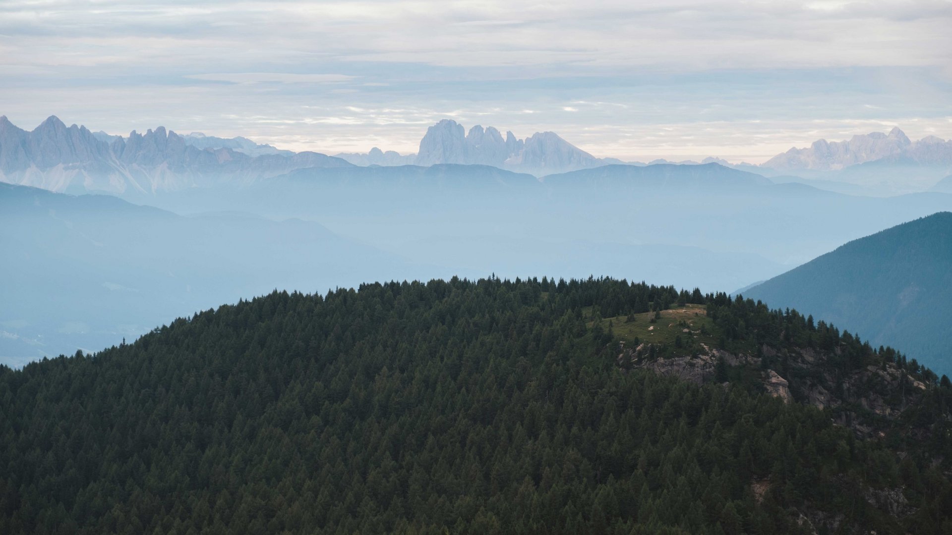 Vals - Jochtal - Steinermandl The picture shows a hilly landscape with densely forested mountains in the foreground. In the background, several mountain ranges are visible, appearing in various shades of blue as they are enveloped by fog or haze, while the sky is cloudy.
