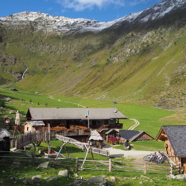 Wieserhütte The picture shows a mountain hut in a green valley surrounded by mountains. In front of the hut there are wooden fences, a small bridge, and several bicycles. The surrounding mountains have occasional snow-capped peaks, and a clear blue sky stretches above.
