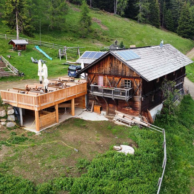 Pertinger Alm The picture shows a rustic wooden mountain cabin with a large terrace and balconies, surrounded by forest and meadows. In front of the cabin are tables and chairs, and solar panels can be seen on the roof.