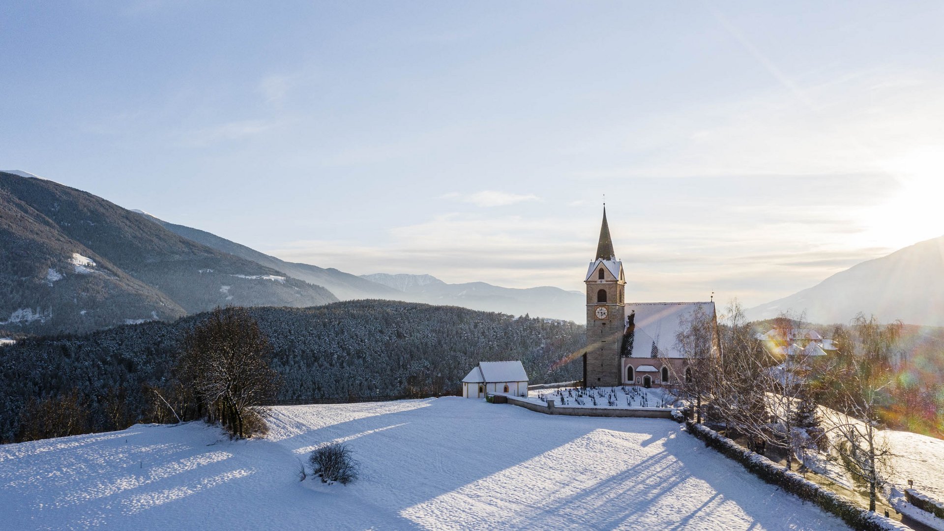 Rodeneck: idylický turistický ráj Obrázek ukazuje kostel s vysokou věží, který stojí v zasněžené krajině. V pozadí jsou zalesněné kopce a hory pod jasnou oblohou, zatímco slunce je nízko na obzoru a vrhá dlouhé stíny.