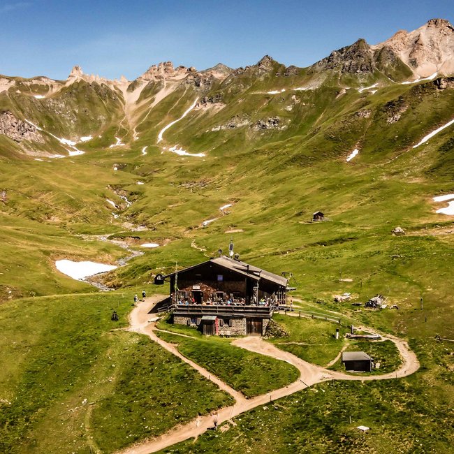 Brixner Hütte Obrázek ukazuje horskou chatku uprostřed zelené horské krajiny s roztroušenými sněhovými políčky. V pozadí se tyčí strmé, skalnaté hory pod jasně modrou oblohou. Malý potok protéká údolím vedle chatky.