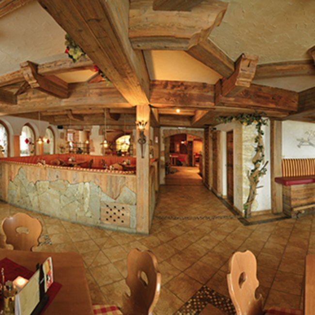 Restaurant Pizzeria Bar Igloo s’Pfandl Obrázek ukazuje interiér rustikální restaurace s dřevěnými trámy na stropě a dřevěným nábytkem. Stoly jsou prostřeny červenými podložkami a na stěnách visí tradiční dekorace jako jelení parohy a rostliny.