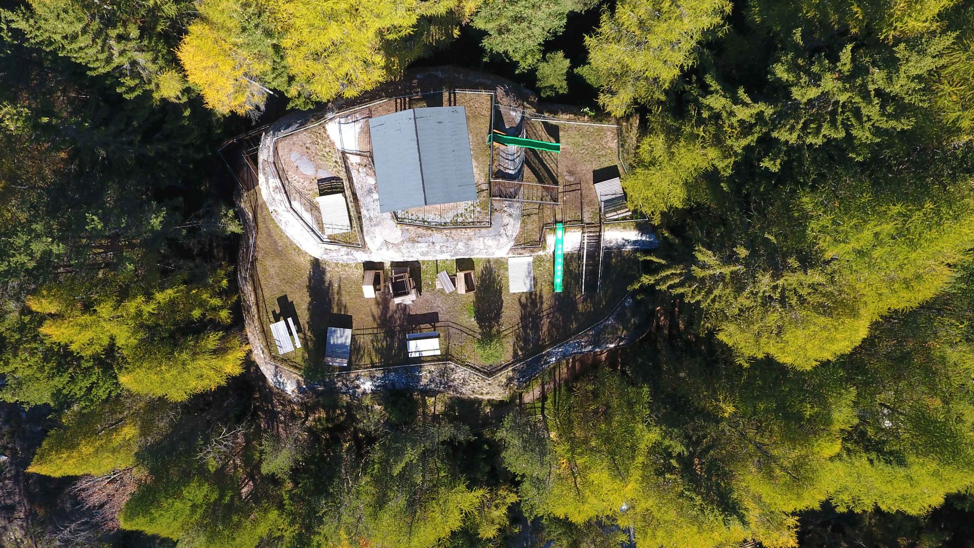 The recreation area in Spinges, South Tyrol The image shows an aerial view of a building located in the middle of a dense forest. Around the building, several benches and tables as well as playground equipment are set up, and the surroundings appear calm and natural.