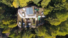 The recreation area in Spinges, South Tyrol The image shows an aerial view of a building located in the middle of a dense forest. Around the building, several benches and tables as well as playground equipment are set up, and the surroundings appear calm and natural.