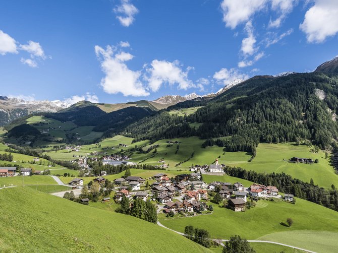 The Alpine hamlet of Spinges The picture shows a picturesque Alpine landscape with a small village surrounded by green meadows and wooded hills. In the background, snow-covered mountains can be seen under a clear, blue sky with some clouds.