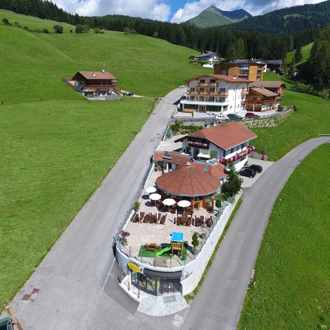 Restaurant Pizzeria Bar Igloo s’Pfandl Obrázek ukazuje letecký snímek několika budov na zelené, kopcovité krajině. Budovy zahrnují restauraci s terasou a bílými slunečníky, několik obytných domů a menší stavby podél asfaltované cesty. V pozadí jsou vidět zalesněné kopce a jasná modrá obloha.