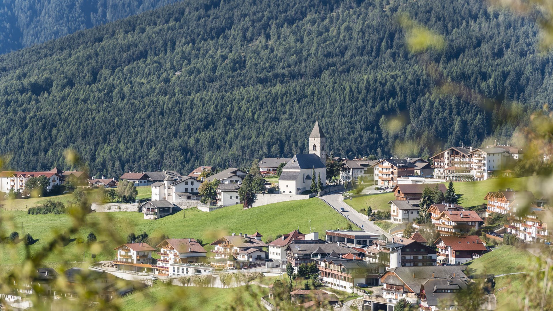 The mountain village of Meransen The picture shows an idyllic village with a central church and a pointed tower, surrounded by residential houses and buildings. The village lies in a green valley, surrounded by dense, forested mountain slopes. The vegetation in the foreground is slightly blurred, which gives the image depth.