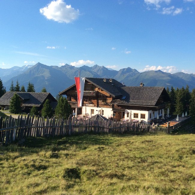 Rastnerhütte Obrázek ukazuje tradiční horský dům s dřevěnou a kamennou konstrukcí, obklopený loukou a dřevěným plotem. V pozadí jsou vidět vysoké zasněžené hory a husté lesy, které tvoří malebnou alpskou krajinu.