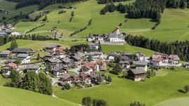 Pfunders: urig und idyllisch Das Bild zeigt ein kleines Dorf in einer grünen, hügeligen Landschaft. Im Zentrum des Dorfes steht eine Kirche mit einem roten Zwiebelturm, umgeben von traditionellen Häusern, und im Hintergrund erstrecken sich saftige Wiesen und dichte Wälder.