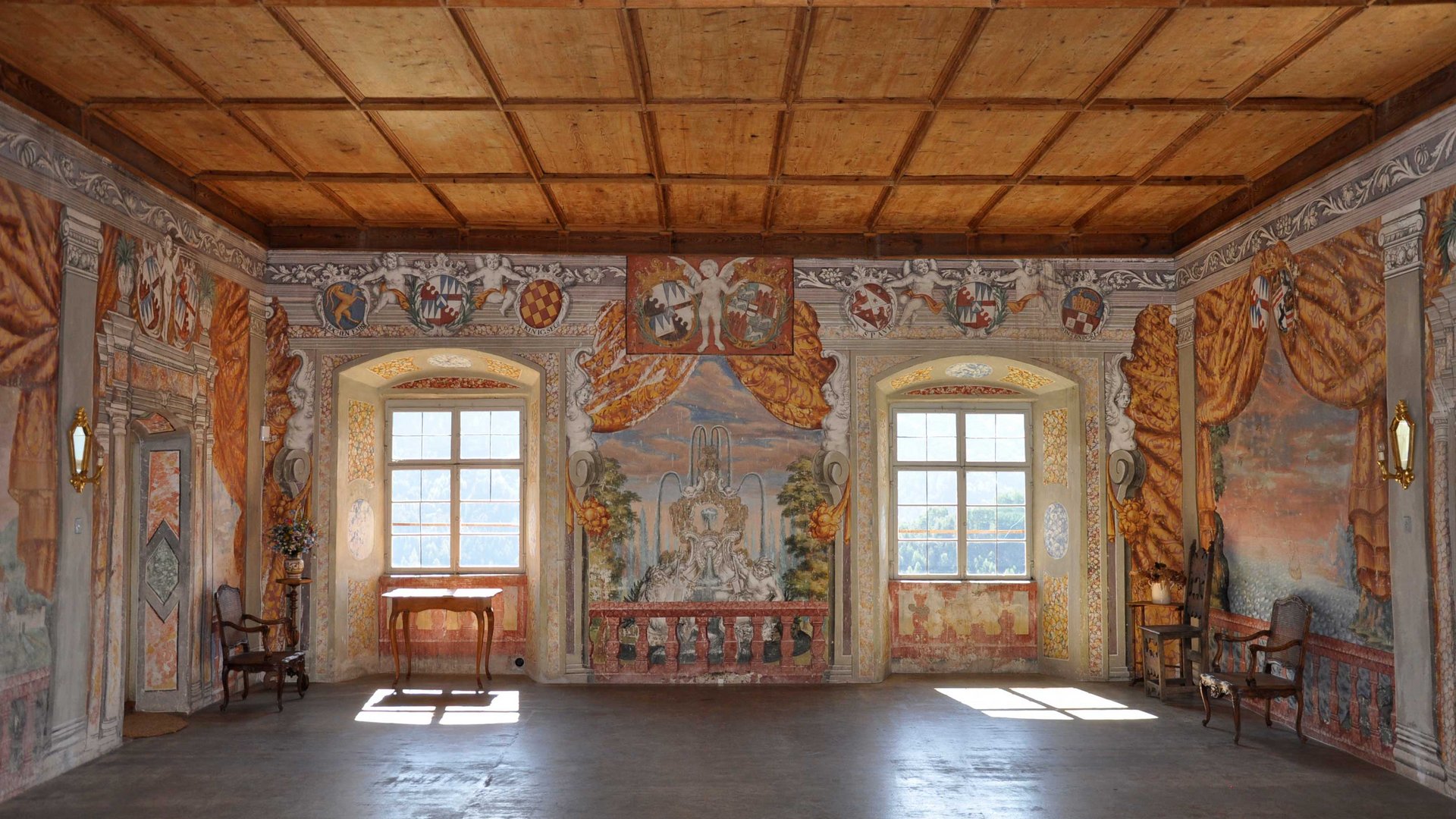 Mächtig und erhaben: Schloss Rodenegg Das Bild zeigt einen prächtigen Raum mit aufwendig bemalten Wänden, die Szenen mit Engeln, Wappen und Landschaften darstellen. Der Raum hat eine Holzbalkendecke, zwei große Fenster, die Licht hereinlassen, und ist mit antiken Möbeln ausgestattet.