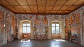 Mächtig und erhaben: Schloss Rodenegg Das Bild zeigt einen prächtigen Raum mit aufwendig bemalten Wänden, die Szenen mit Engeln, Wappen und Landschaften darstellen. Der Raum hat eine Holzbalkendecke, zwei große Fenster, die Licht hereinlassen, und ist mit antiken Möbeln ausgestattet.