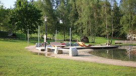 Het Aktivpark Terenten De afbeelding toont een rustige park met een kleine vijver, waarin houten ligstoelen op een zanderig oppervlak staan. Op de achtergrond staan meerdere bomen en de omgeving wordt gekenmerkt door veel groen en natuur.