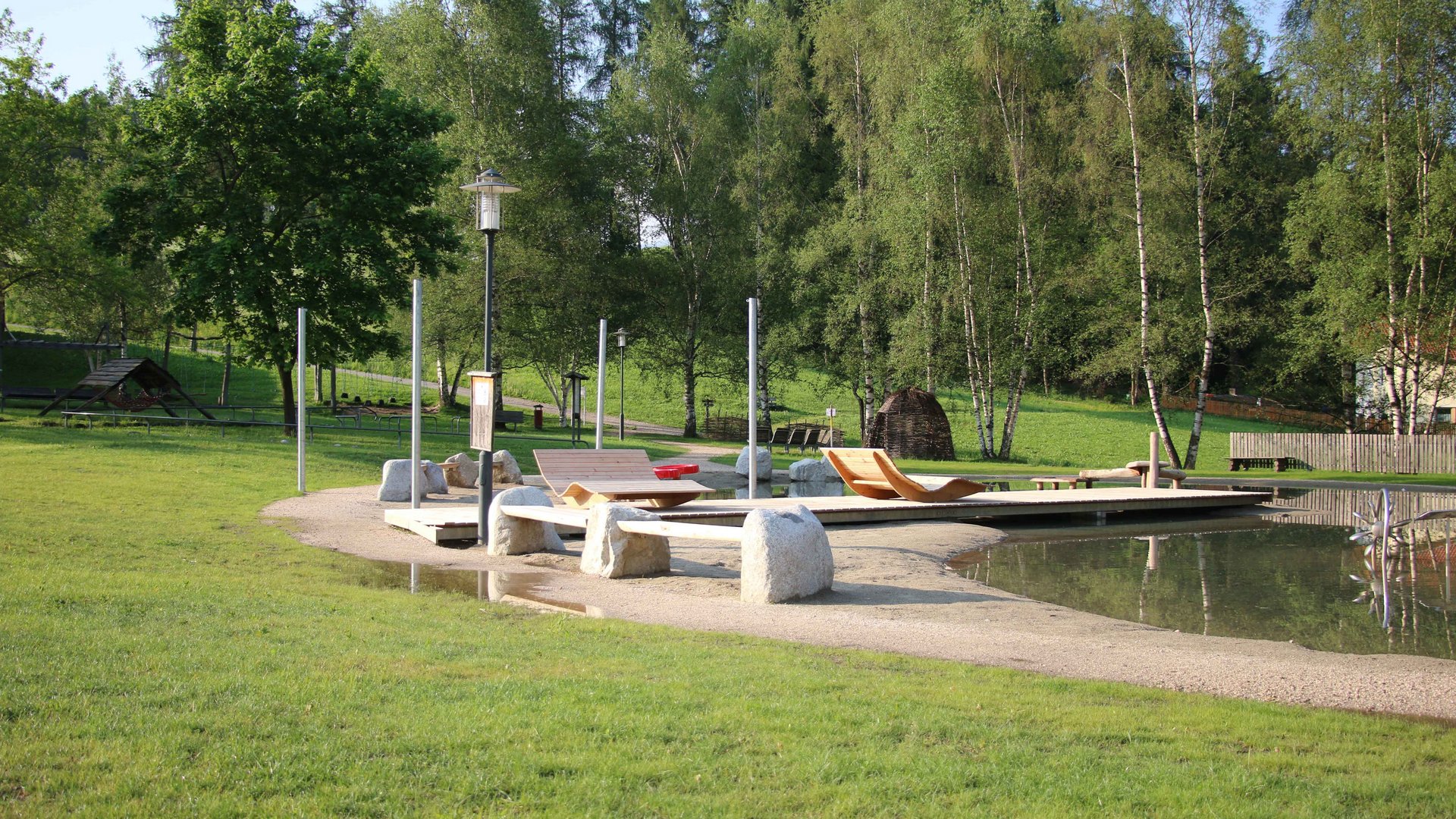 Het Aktivpark Terenten De afbeelding toont een rustige park met een kleine vijver, waarin houten ligstoelen op een zanderig oppervlak staan. Op de achtergrond staan meerdere bomen en de omgeving wordt gekenmerkt door veel groen en natuur.