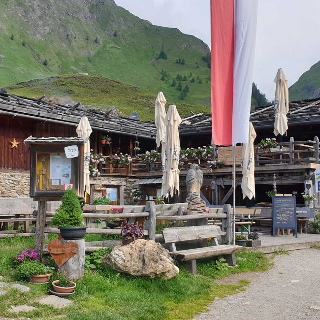 Zingerle Hütte De foto toont een rustieke berghut met houten en stenen muren, omgeven door groene heuvels. Voor de hut staan meerdere houten banken en tafels, deels met gesloten parasols, en er zijn veel kleurrijke bloemen evenals een houtsnede. Op de voorgrond wappert een grote rood-witte vlag.