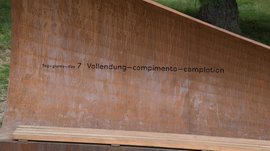 Der Schöpfungsweg in Rodeneck Das Bild zeigt eine metallene Bank oder Skulptur, auf der der Schriftzug "Tag 7 – Vollendung" in mehreren Sprachen eingraviert ist. Die Struktur wirkt rostfarben und steht im Freien, vermutlich in einem naturbelassenen Umfeld.