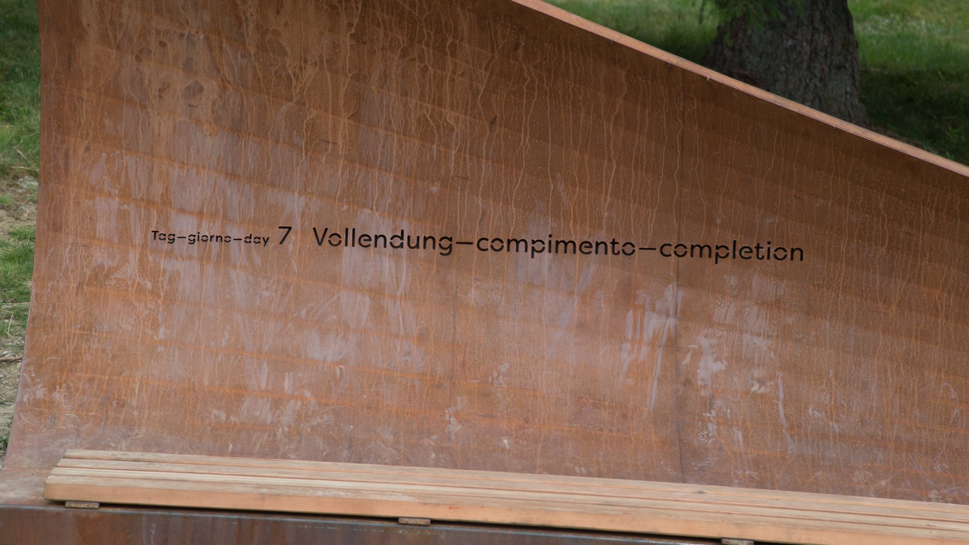 Der Schöpfungsweg in Rodeneck Das Bild zeigt eine metallene Bank oder Skulptur, auf der der Schriftzug "Tag 7 – Vollendung" in mehreren Sprachen eingraviert ist. Die Struktur wirkt rostfarben und steht im Freien, vermutlich in einem naturbelassenen Umfeld.