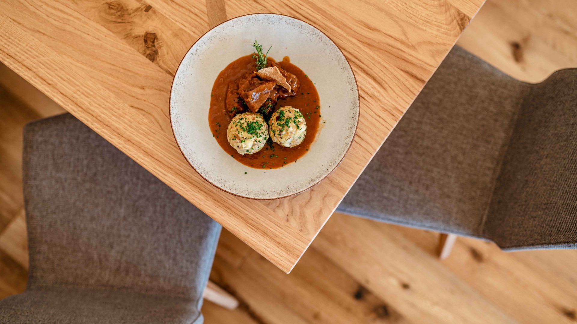 Hütten in Meransen, Vals und Co. Auf dem Bild ist ein modern angerichteter Teller mit zwei Speckknödeln und Gulasch auf einem Holztisch, gesehen aus der Vogelperspektive, mit zwei leeren Stühlen im Hintergrund.