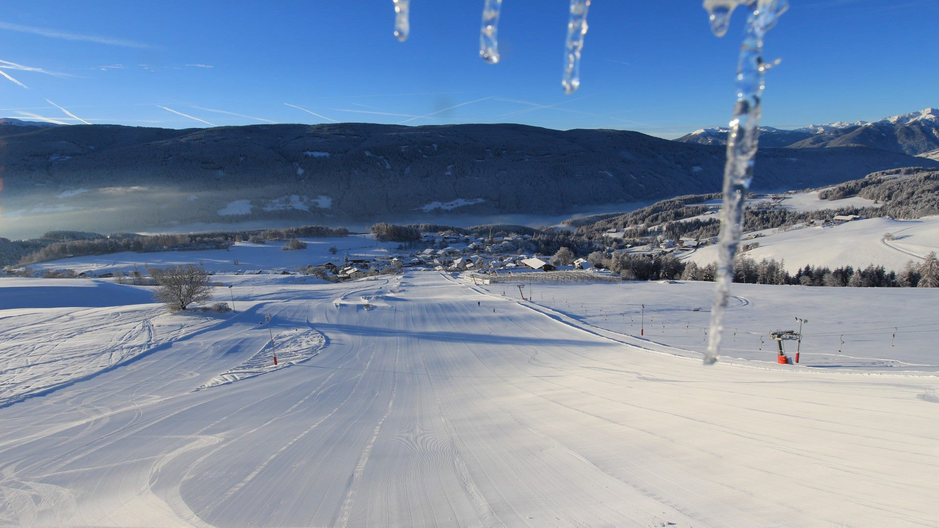 Skivakantie met kinderen in Zuid-Tirol/Terenten De afbeelding toont een met sneeuw bedekt dorp met huizen en een kerk temidden van een winterse berglandschap. Op de achtergrond trekken mistbanken door het dal, terwijl de lucht helder en blauw is.